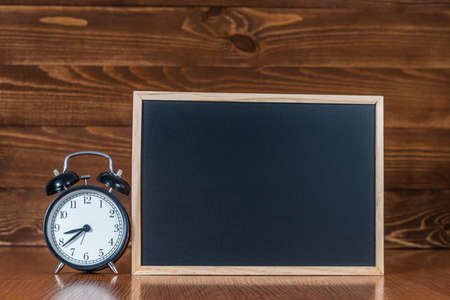 A blackboard with space for text and an alarm clock on a wooden background. Concept start of the school year and sale of school supplies. Home education and learning. Mock up back to schoolの写真素材