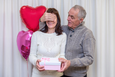 Happy couple seniors celebrate Valentines Day. Man closes the womans eyes and gives her gift. Romantic relationships, love and dating in retirement. Lifestyle of elderly people. Mature pensioners.の写真素材