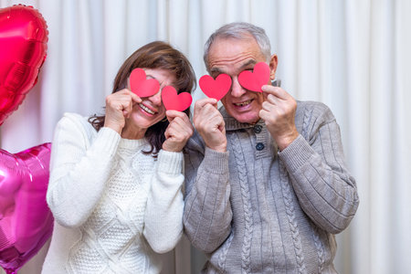 Crazy couple seniors celebrate Valentines Day. Man and woman cover eyes with red valentine cards and smile. Romantic relationships, love and dating in retirement. Lifestyle of elderly people.の写真素材