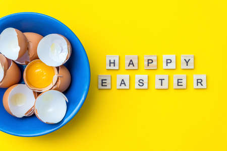 bright creative postcard with happy Easter text. Raw eggs, yolk and shell in a blue plate on a yellow background. The concept of preparing for the holiday. Festive omelet or scrambled eggs.の写真素材