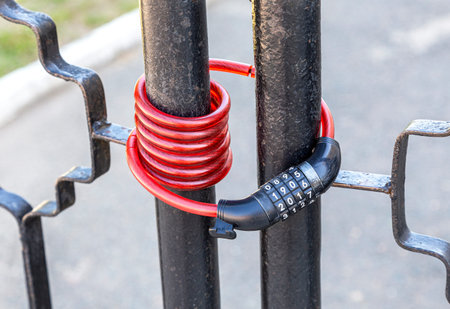 Flexible red bicycle lock with the code set hangs on metal gates, close up. Security and safety...
