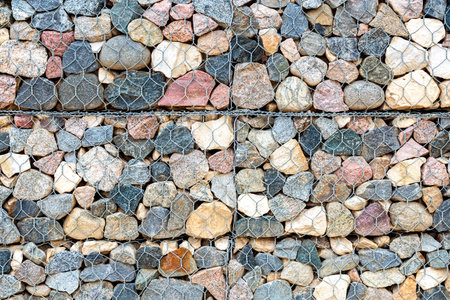 Stone wall with metal grid and rough stones as background. Rock texture in metal grid. Gabion net...