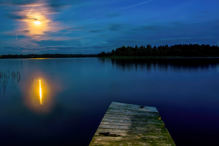 Night view of the lake with an old wooden pier and the moon reflecting on the surface of the waterの写真素材