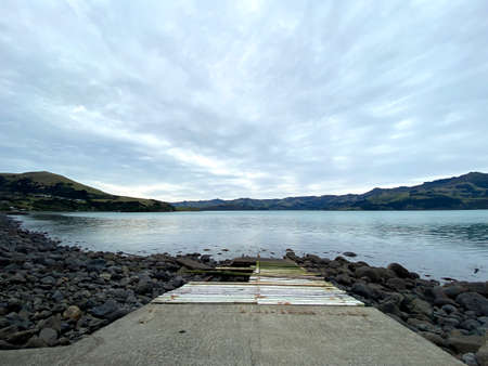 Bot Ramp, Wainui, Canterbury, New Zealand. Cloudy day, dramatic shot of an old boat slip going out on a rock shore to a beautiful calm inlet. Abandoned old wooden boat ramp.の写真素材