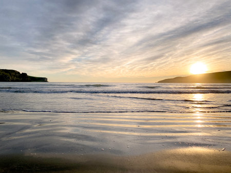 Okains Bay, Canterbury, New Zealand. Sunrise over beautiful sandy beach with hills and gentle coastline. Small seaside settlement near Akaroa. Golden sand with dramatic cloud formations overhead.の写真素材