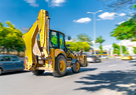 The excavator drives or goes fast on the street with heavy traffic with blue sky and green trees in...