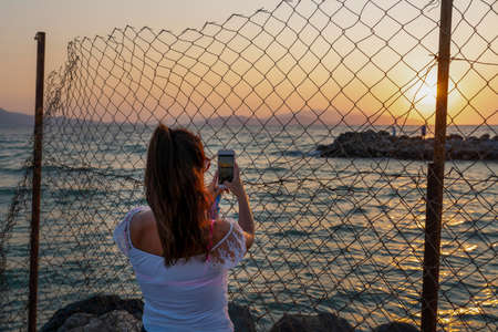 Back rear view of woman taking photo of sunset with mobile phone from behind fenceの写真素材
