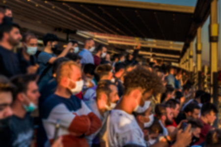 Unfocused blurred face masked spectators watching the match standing in the stadium with a mobile phone in their hands.のeditorial素材