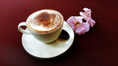 Hot cappuccino or latte coffee. A cup of coffee and pink trumpet flower on the table with red-brown...