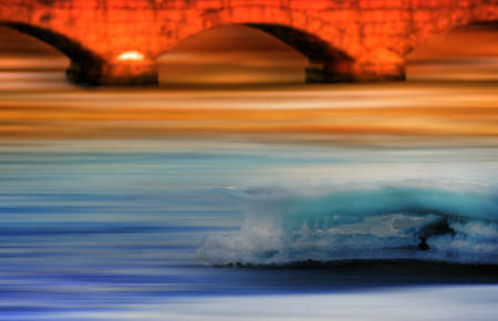 River at spring with abstract motion, reflecting colorful sunset, old bridge in backgroundの写真素材