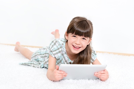 Joyful pretty little girl laughing as she lies on her stomach on a white carpet playing with a modern digital tablet in a white room の写真素材