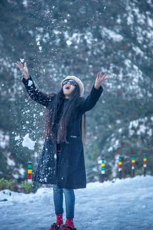 Portrait of an young and attractive Indian woman wearing woolen cap and jacket enjoying snowfall and feeling amazed in a snowy eveningの写真素材