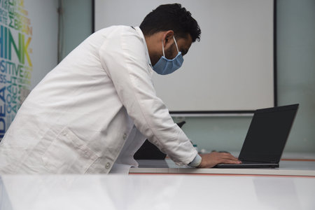 Asian doctor in white coat wearing mask and stethoscope working on laptop at clinic. Health care,...