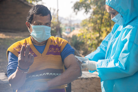 Female Nurse with PPE kit protective mask and gloves preparing a male patient's arm for vaccination in Rural village of India. Healthcare workers giving vaccine to a people in pandemic.の写真素材