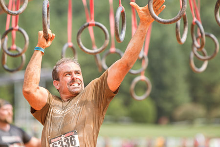Conyers, GA, USA - August 22, 2015:  A man grimaces as he swings from rings suspended from wood beams to keep him out of muddy water at the Rugged Maniac Obstacle Course race in Conyers, GA.のeditorial素材
