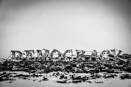 Barbed wire envelops charred and battered wooden letters spelling out the word "Democracy," suggesting democracy is under attack.の写真素材