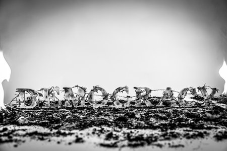 Black and white shot of barbed wire enveloping charred and battered wooden letters spelling out the word "Democracy," suggesting democracy is under attack.の写真素材