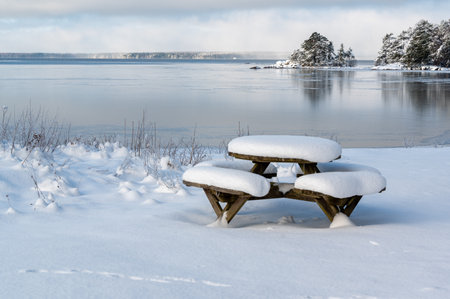 Table with benches covered in snow infront of lakeの写真素材