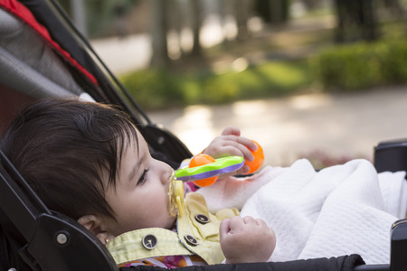 Little baby resting at baby carriage and playing with toys, yellow dummy inside her mouth. outdoor...