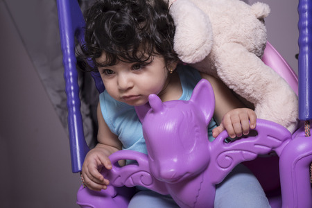 Portrait of a sleepy little baby girl on unicorn swing with teddy bear doll on herの写真素材