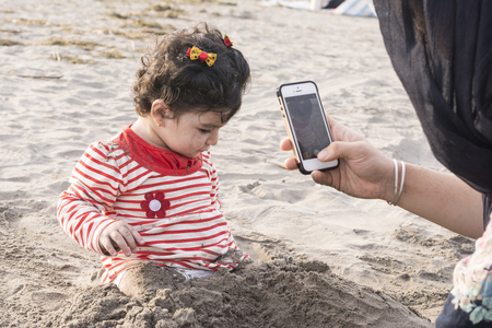Little kid playing at beach with sand and her mother taking photos by smartphoneの写真素材