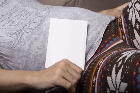 Pregnant woman holding a small blank catalogue in front of belly resting on sofaの写真素材