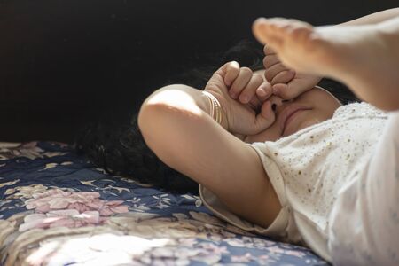 Little girl lied down on bed and touching her eyes by hands, home interior window natural sunlight...