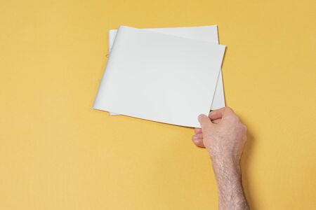Male hand touching a catalog with blank cover on yellow ground, mock-up series template ready for...