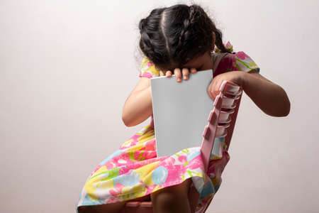 Shy little girl hiding her head behind a story book with blank cover in front of her body, editable mock-up series template ready for your design. cover selection path included.の写真素材