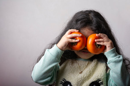 Little girl holding two persimmons in fornt of eyes, covers her face.の写真素材