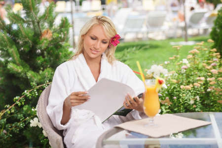Carefree beauty. Attractive young women in bathrobe reading the magazine while sitting at the table with a cocktail on itの写真素材