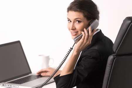 Talking at phone. Confident middle-aged businesswoman sitting at her working place and looking over shoulderの写真素材