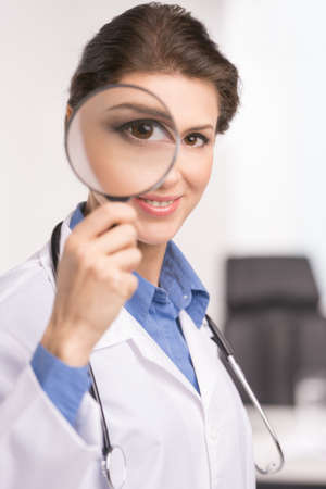 Doctor with magnifying glass. Confident middle-aged female doctor holding a magnifying glass in front of her eyeの写真素材