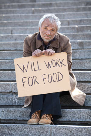 Will work for food. Depressed man in dirty wear sitting on stairs and holding poster