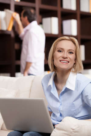 Happy woman. Cheerful middle aged woman working at the computer and smiling while her husband choosing a book to read at the backgroundの写真素材