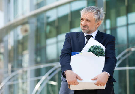 Fired. Depressed senior man in formalwear holding box with his work stuff and looking awayの写真素材