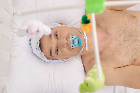 Big baby. Top view of infant adult man with pacifier looking at camera while lying on the baby bedの写真素材
