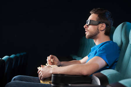 Men at cinema. Side view of excited young men in glasses watching movie at the cinema and eating popcornの写真素材