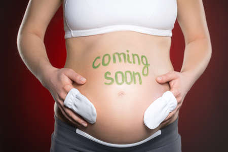 Pregnancy. Close-up of pregnant woman touching her abdomen while standing against red backgroundの写真素材
