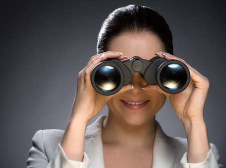 Look in future. Portrait of confident middle-aged woman in formalwear looking through binoculars while isolated on greyの写真素材