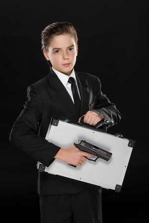 Little agent. Cheerful little boy in formalwear holding a suitcase and handgun while standing against black backgroundの写真素材