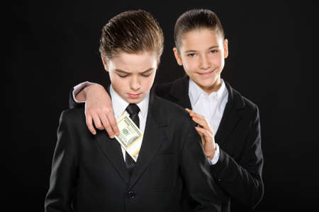 Business kids. Cheerful little girl in formalwear putting money to little boy pocket and smiling while standing against black backgroundの写真素材