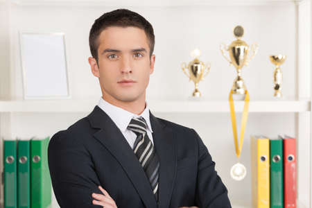 Confident businessman. Confident young businessman standing in front of his business trophies and holding his arms crossedの写真素材