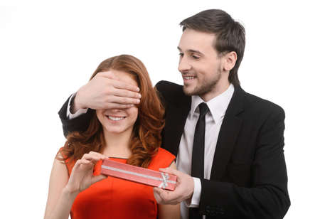 Young men in full suit closing his girlfriends eyes with the hand and presenting a gift box against white backgroundの写真素材