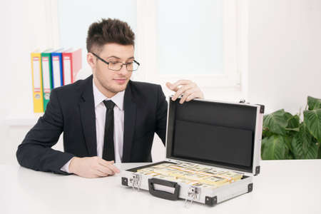 Wealthy businessman. Handsome young businessmen in glasses looking at the suitcase full of moneyの写真素材
