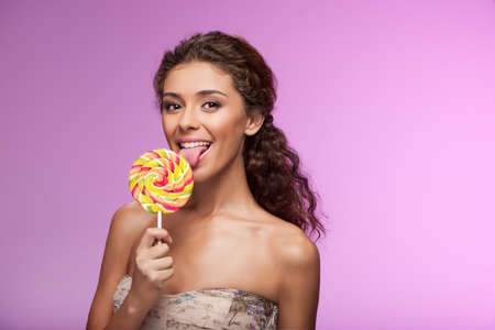 Eating a lollipop. Beautiful young women eating lollipop while standing isolated on colored backgroundの写真素材