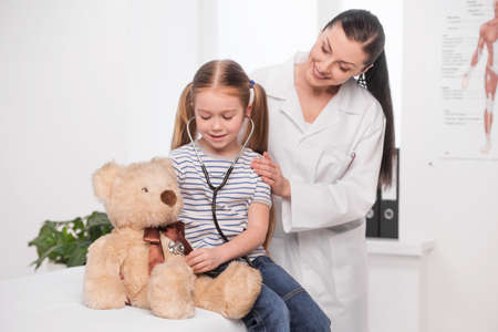 Doctor and little girl. Cheerful young doctor in lab coat examining little girl with stethoscopeの写真素材