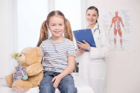 Child on medical exam. Cheerful little girl looking a camera and smiling while doctor standing on the background and holding clipboardの写真素材