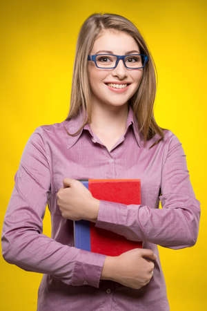 Cheerful female student. Smiling young women holding books in her hand while standing against yellow backgroundの写真素材