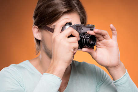Man with camera. Confident young man holding camera in his hands and focusing at you while isolated on colored backgroundの写真素材